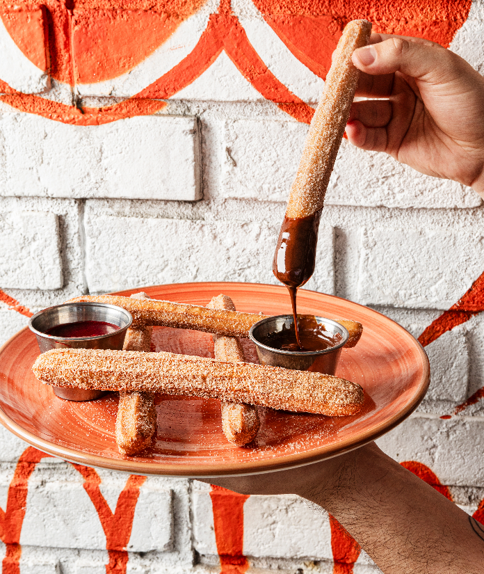Hand dipping a churro into melted chocolate on a plate with more churros and dipping sauces.