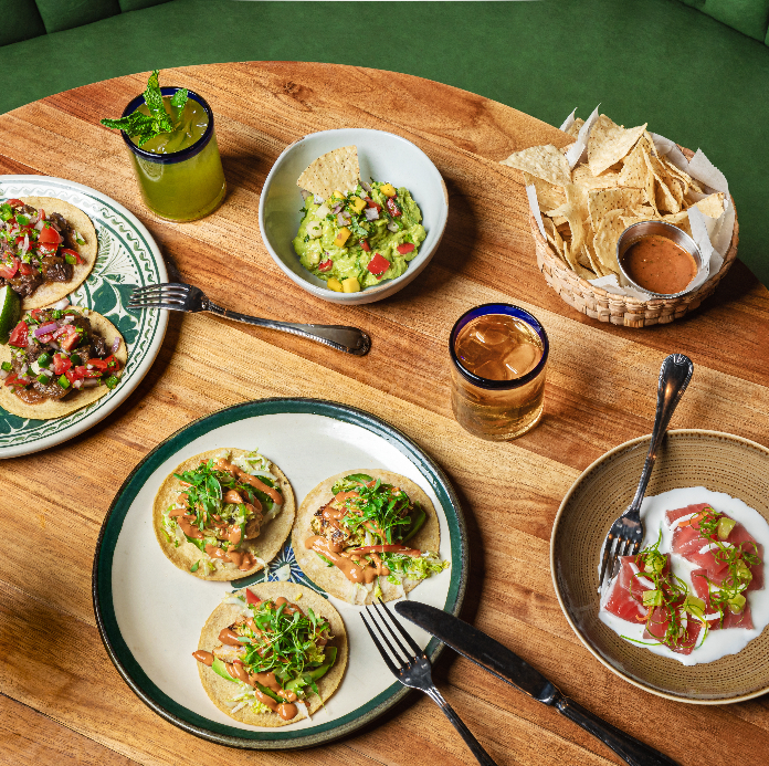 A wooden table set with tacos, guacamole, chips, and drinks, showcasing a Mexican meal setup.