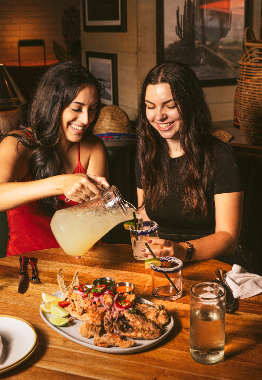 Two women sharing a meal and drinks at a wooden table, one pouring a beverage from a pitcher into a glass