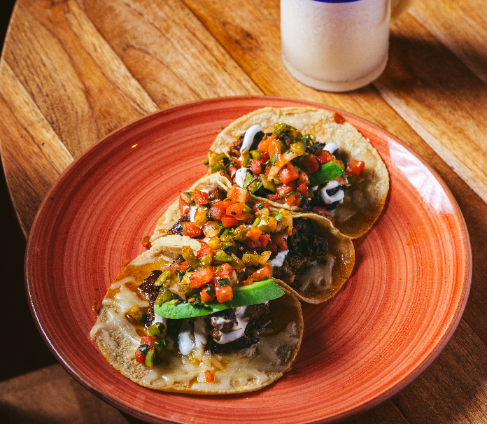 Three street tacos on corn tortillas topped with pico de gallo, avocado, and crema, served on a red plate on a wooden surface.