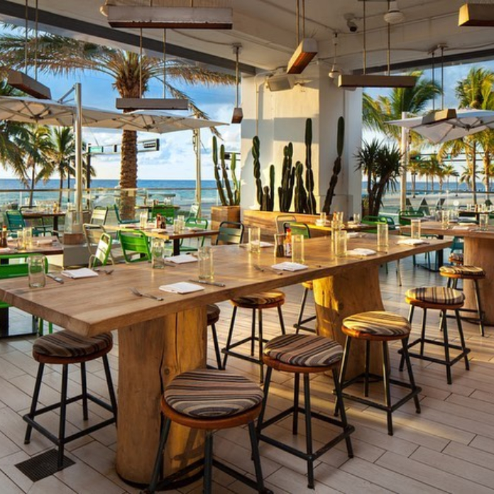 Modern outdoor restaurant with wooden tables, bar stools, cacti, palm trees, and ocean view.