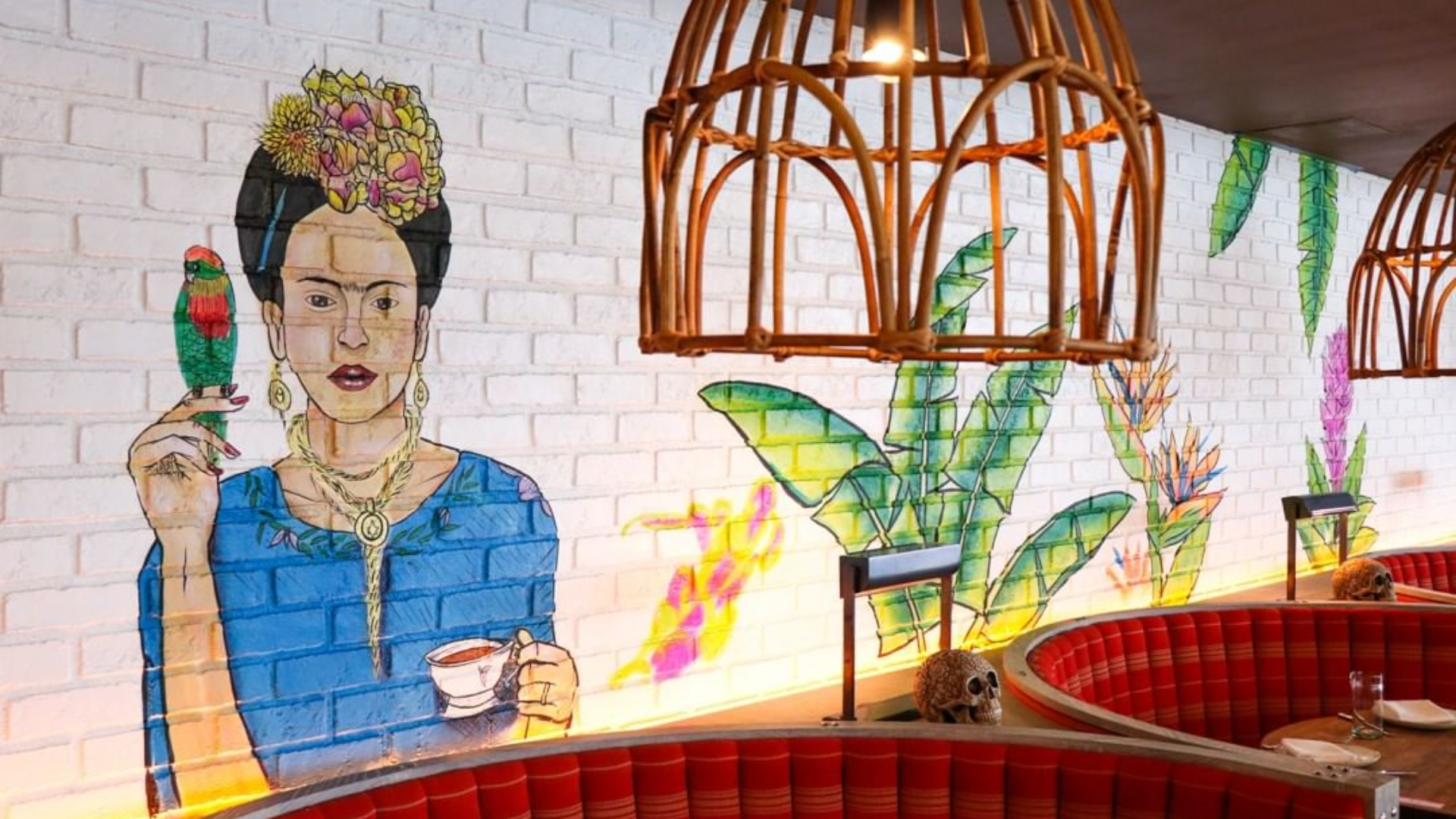 Frida Kahlo mural, birdcage, green plants, red seating, skull decor at a restaurant.