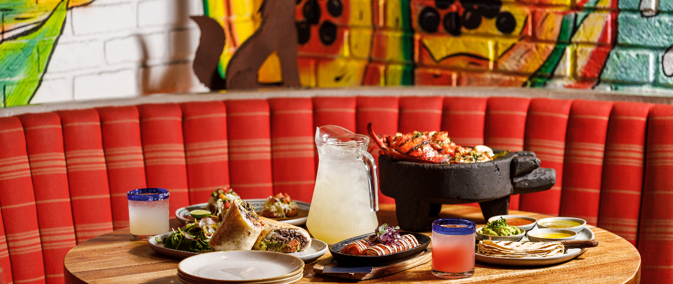 A spread of Mexican dishes on a wooden table, including a molcajete, tacos, and colorful drinks, set against a red booth and vibrant mural.